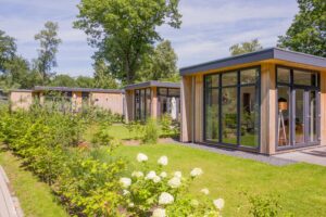Multiple sleek, flat-roofed buildings with natural wood cladding and floor-to-ceiling glass windows, located in a well-maintained green space with plantings and trees. A year-round garden room.