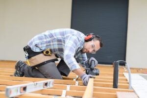 Person engaged in constructing a wooden deck frame, kneeling and using a power tool to fasten beams, while wearing protective gear. This deck is standing on screw piles, which are great for both professional and diy deck projects.