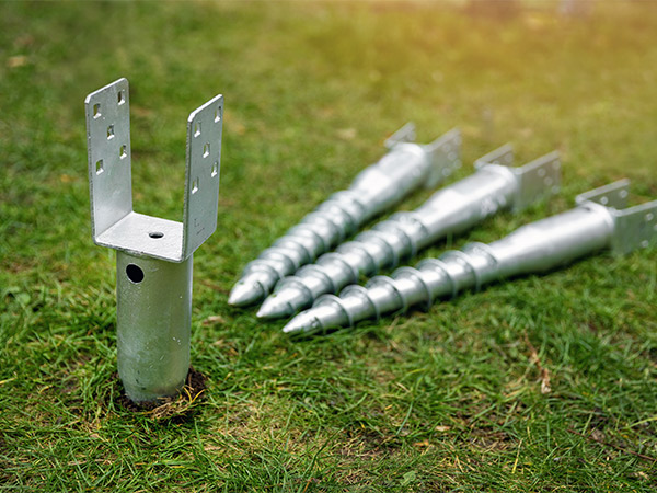 A close-up outdoor shot of four GroundPlug® screw piles and a U-bracket on green grass. One screw pile is upright, with the U-bracket attached to its top, showing a metal post with a flat base and a U-shaped bracket with holes for attachment. Three other screw piles are laid horizontally in a row behind it, each showing a helical flange design.