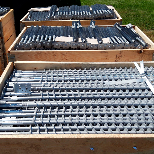 Three wooden pallets are stacked outdoors, filled with GroundPlug® Twister screw piles. The pallets are arranged in a tiered fashion, with the top pallet showing a variety of black components, possibly sleeves or protective covers, alongside metal elements.
