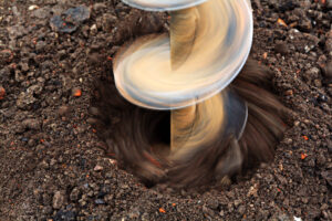 A close-up, overhead shot of a metal auger drilling into dark, damp earth. The auger is centered in the frame, and its spiral shape is clearly visible as it rotates. The earth is dark brown with visible clumps and small pieces of debris. The motion of the auger is blurred, indicating its rotation. The scene suggests the process of digging a hole for a post or other construction purpose.