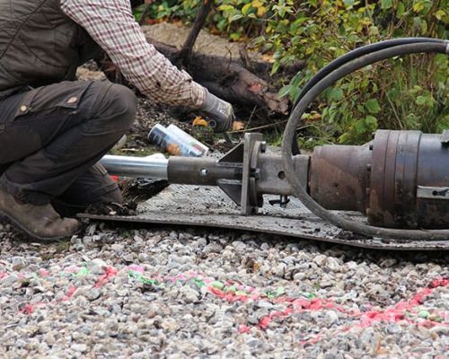 Construction worker preparing or connecting a heavy hydraulic drive head to a foundation element on a gravel surface.