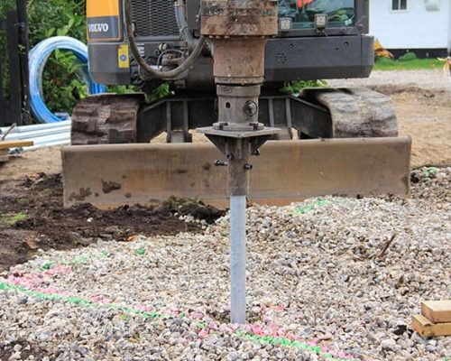 Excavator installing a ground screw foundation element into a gravel bed marked with paint.