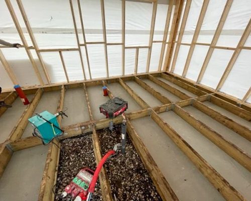 Floor system under construction inside a wood-framed building, showing bays between joists filled with gravel and smooth concrete, with red and grey plumbing pipes, red conduit, and power tools visible.