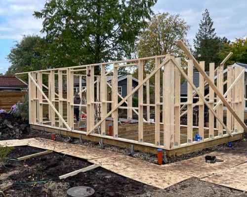 Framing stage of a building project, showing erected wood stud walls, floor system on piers, and initial roof structure assembly.
