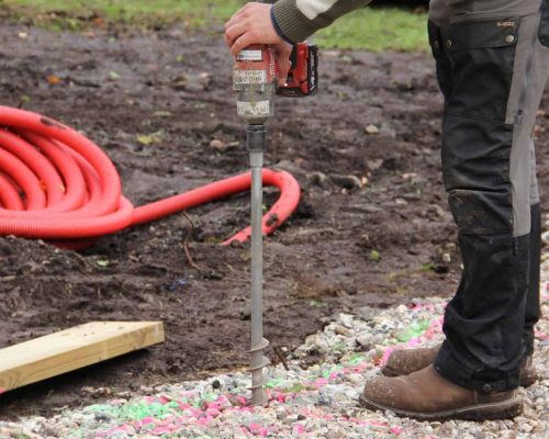 Installing a helical ground screw foundation using a handheld cordless power driver on a prepared gravel base, with orange conduit lying in the background.