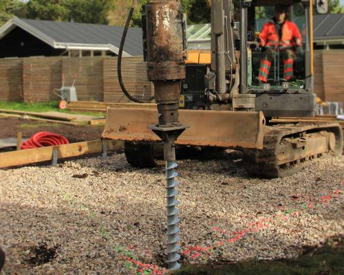 Excavator operated by a person in an orange jacket installs a ground screw on a gravel construction site.